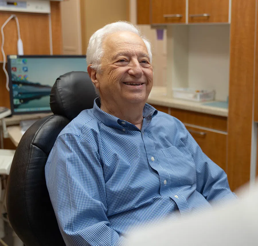 Elderly man smiling in a blue checkered shirt sitting in a comfortable chair in a home office.