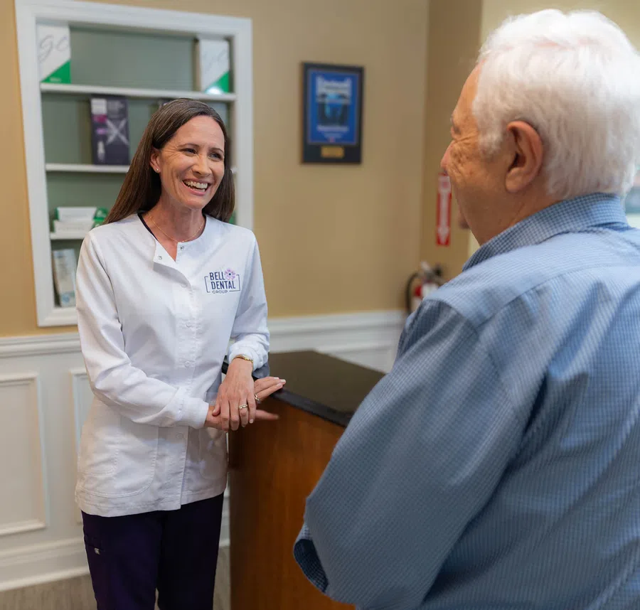 Smiling female dental professional in white coat talking to elderly male patient at dental office reception.