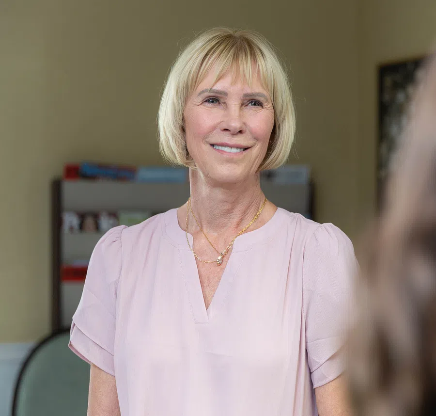 Smiling middle-aged woman with blonde bob haircut wearing a light pink blouse and gold necklace indoors.