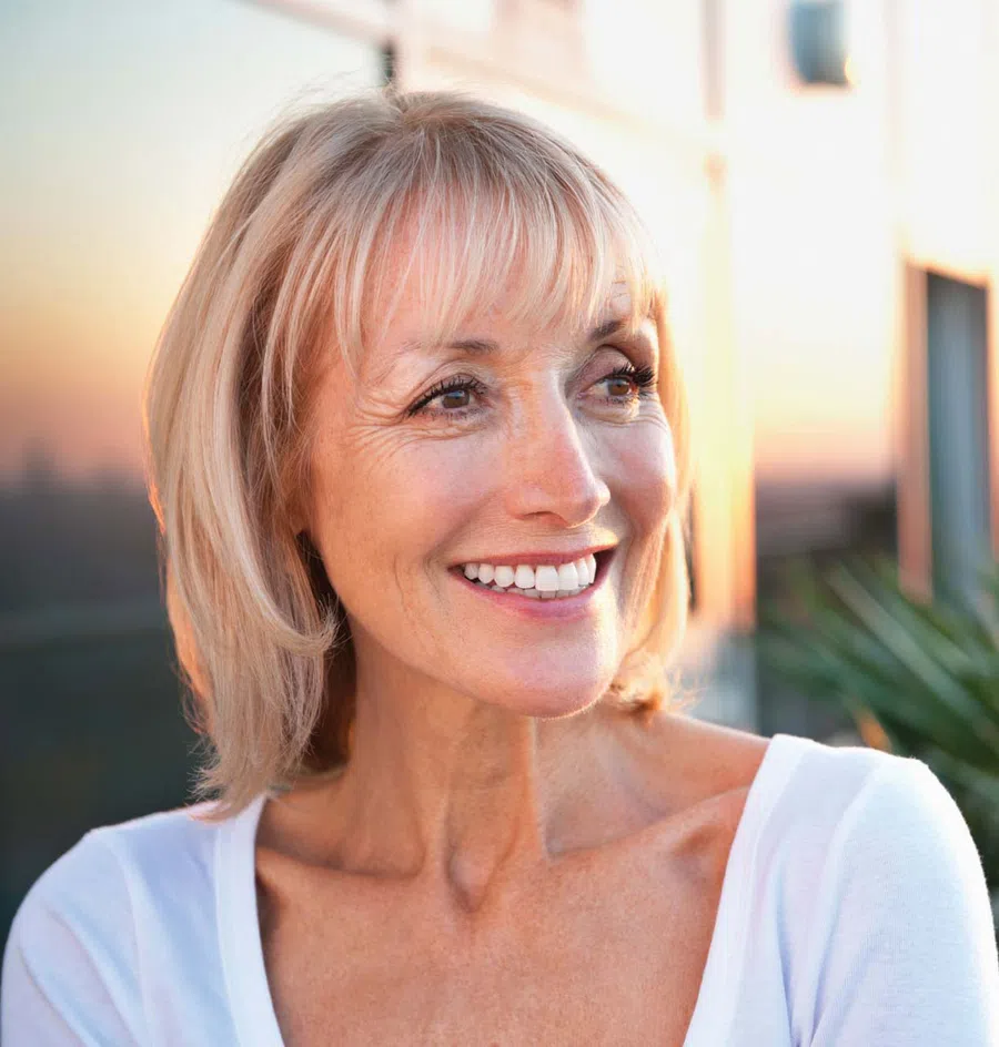Smiling middle-aged woman with short blonde hair in white top, outdoor setting during warm sunset light.