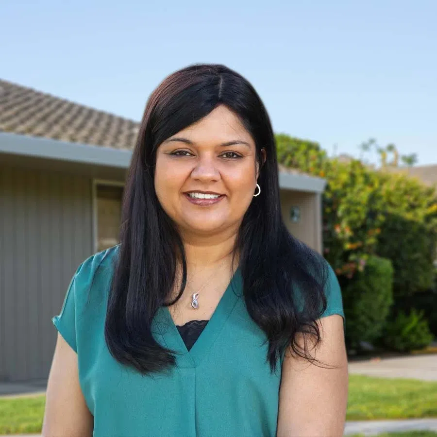 Smiling woman with long black hair wearing teal top standing outdoors in front of a house with green shrubs.