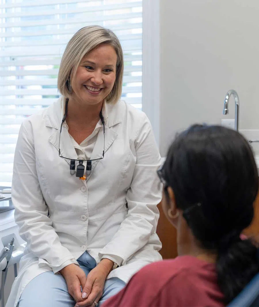 Female dentist smiling and talking to patient in dental office with bright window blinds in background