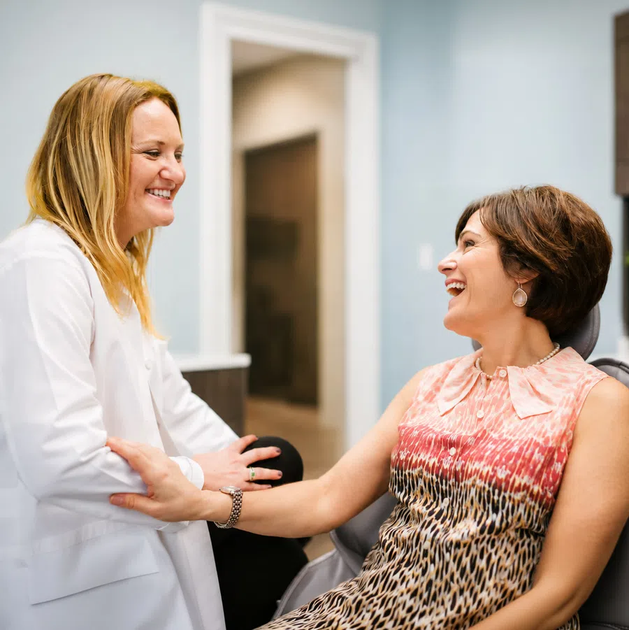 Smiling female doctor in white coat holding hands and talking with happy patient in clinic room.