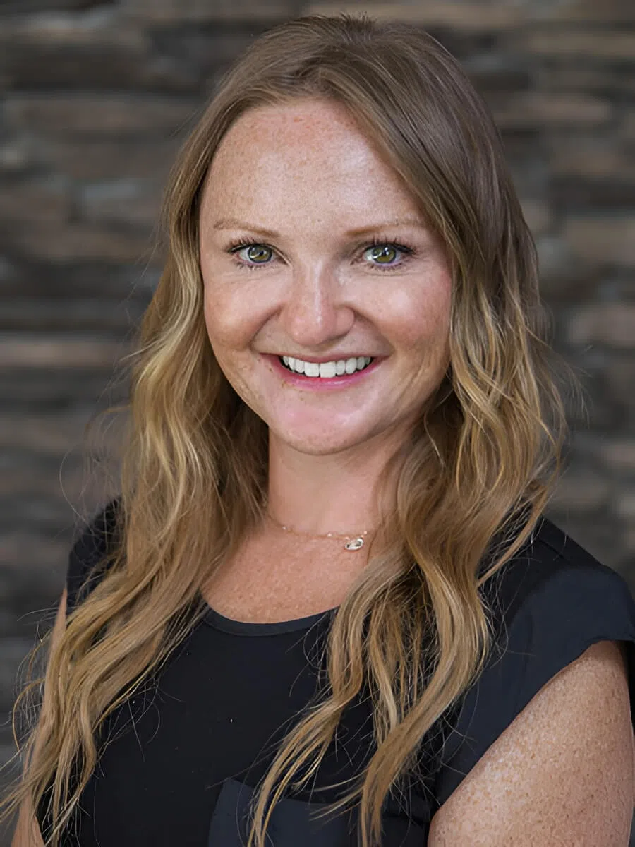 Smiling woman with long wavy blonde hair and freckles wearing a black shirt against a stone wall background.
