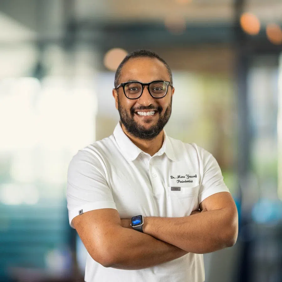 Smiling male dentist with glasses in white polo shirt standing with arms crossed in bright dental clinic.