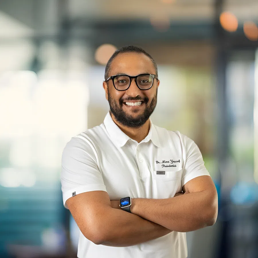 Smiling male dentist with glasses in white polo shirt standing with arms crossed in bright dental clinic.