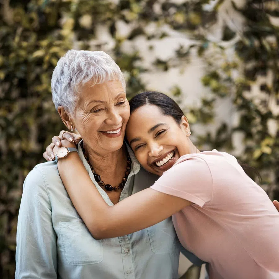 Smiling middle-aged woman and young woman standing close outdoors with greenery in the background