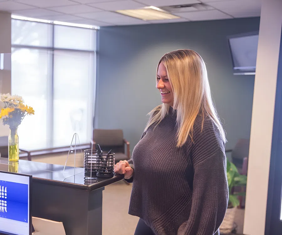 Smiling woman standing at a reception desk in an office with flowers and computer screen visible