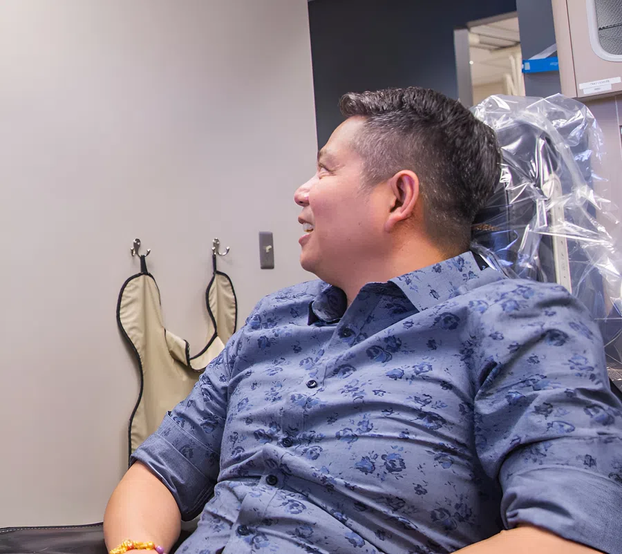 Man in a blue floral shirt sitting and smiling in a medical or dental office with protective aprons hanging on the wall.