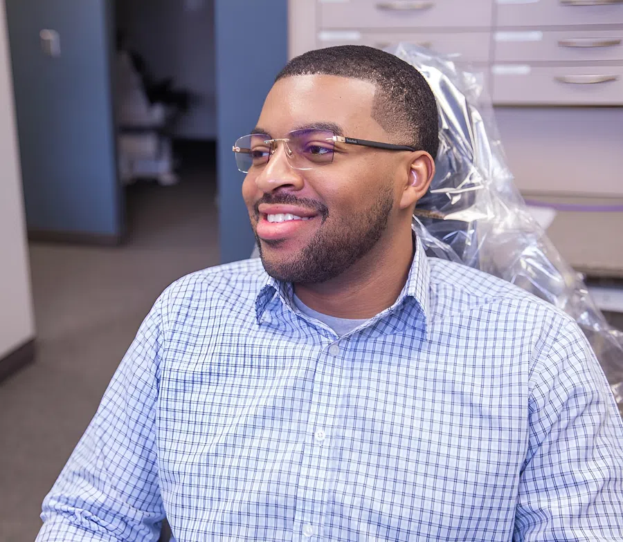 Smiling man wearing glasses and checkered shirt sitting in a dental chair in a clinic environment.