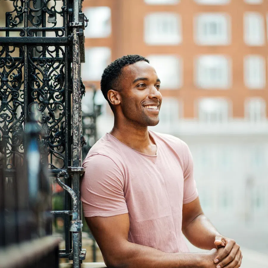Smiling man in a pink t-shirt leaning against decorative black iron gate in urban setting.