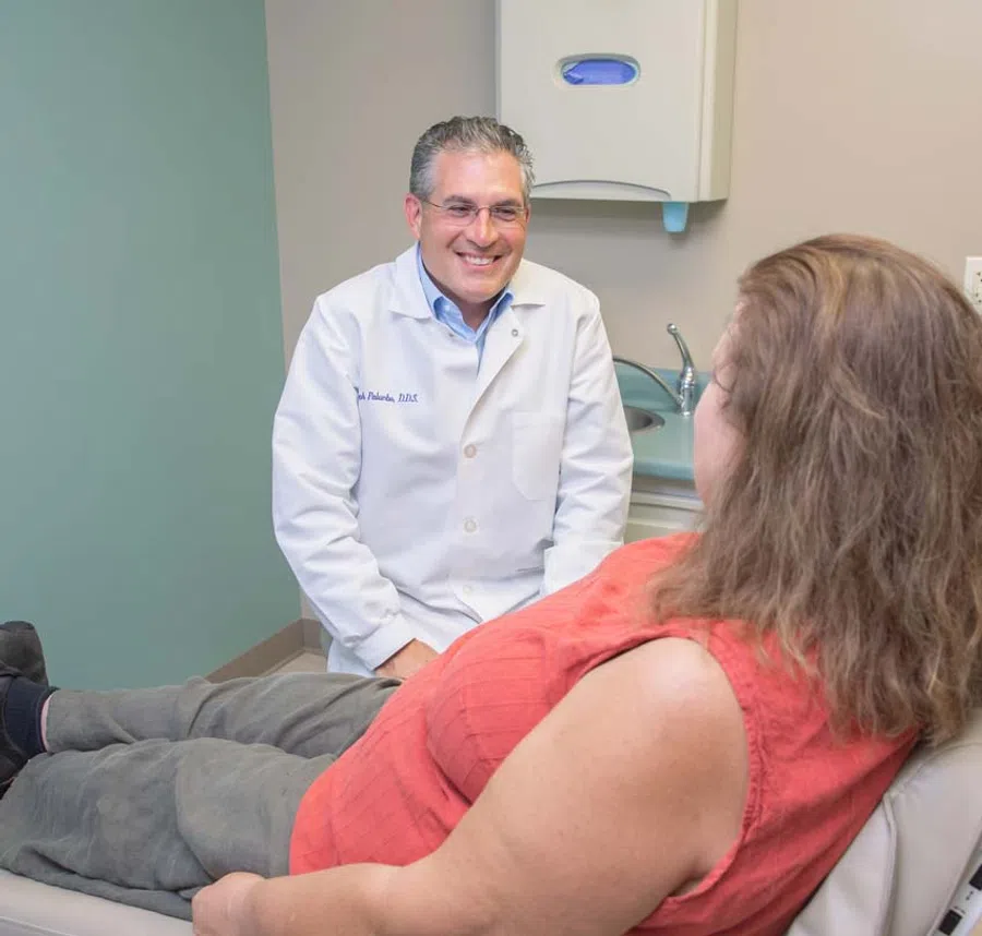 Smiling dentist in white coat consulting female patient reclining in dental office chair during appointment.