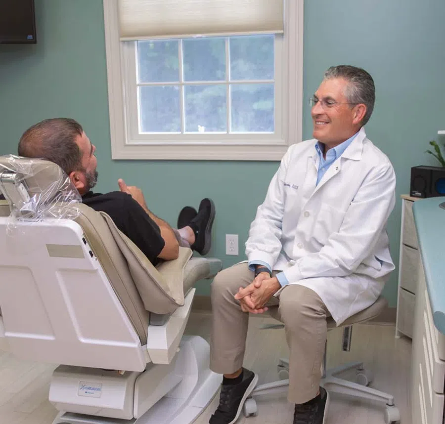 Dentist in white coat consulting a male patient reclining in a dental chair in a bright clinic room.
