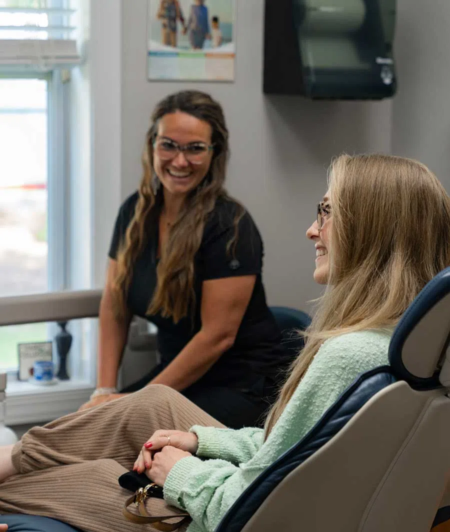 Smiling female patient sits in dental chair talking with a friendly dental hygienist wearing glasses indoors.