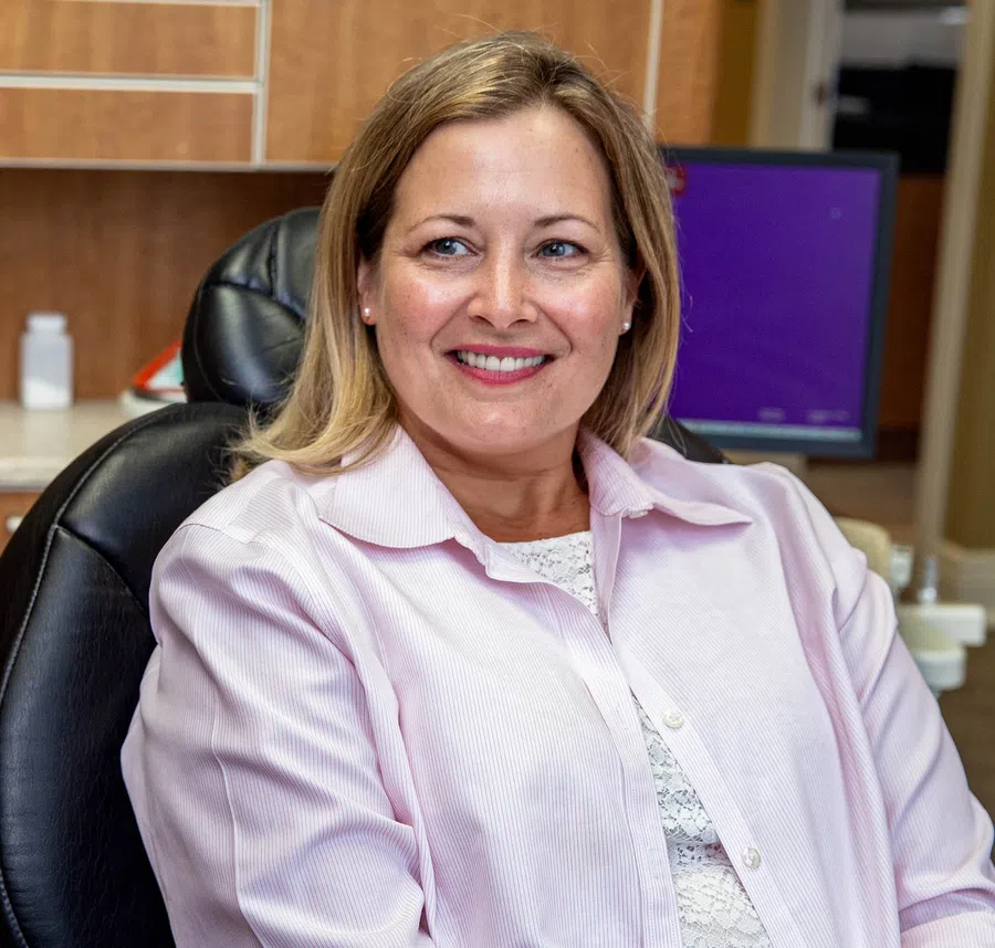Smiling woman with blonde hair wearing a light pink shirt sitting on a black chair indoors.