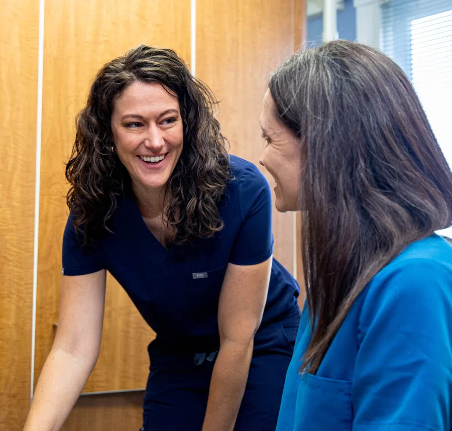 Two female healthcare professionals smiling and interacting in a medical office setting with wooden cabinets.