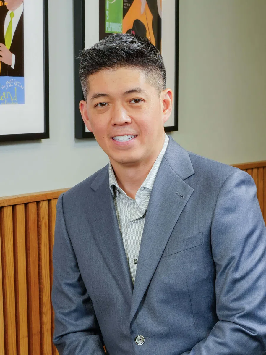 Professional man in gray suit sitting indoors with framed artwork and wooden paneling in background