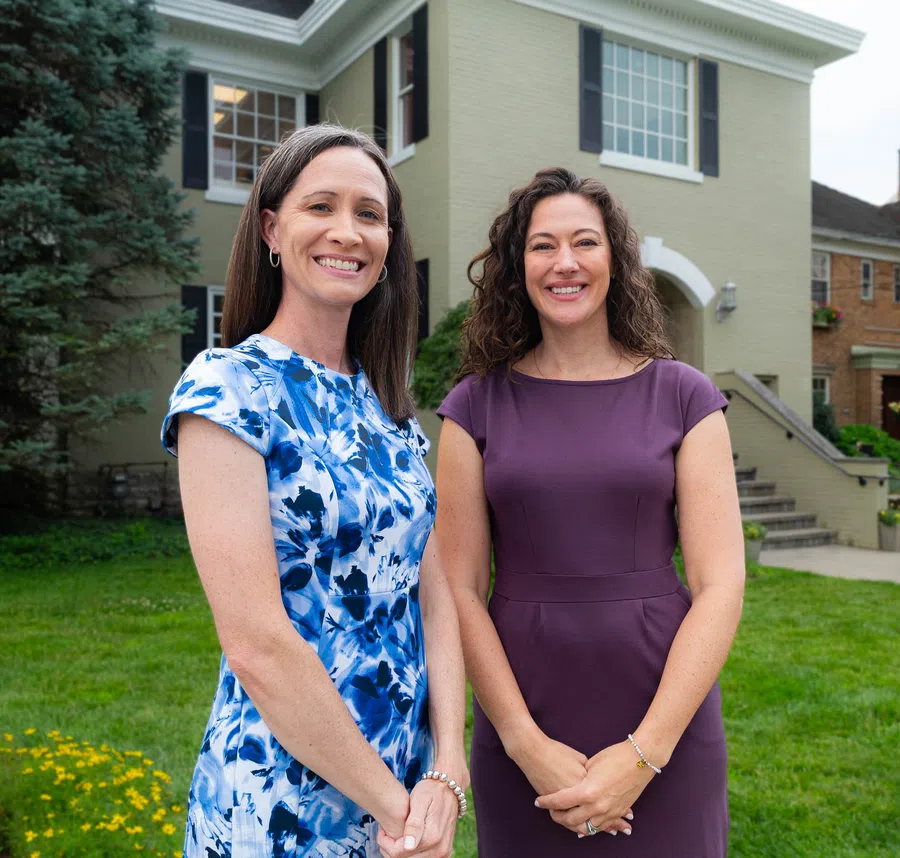 Two smiling women standing outdoors in front of a house, one in a blue floral dress and the other in a purple dress.