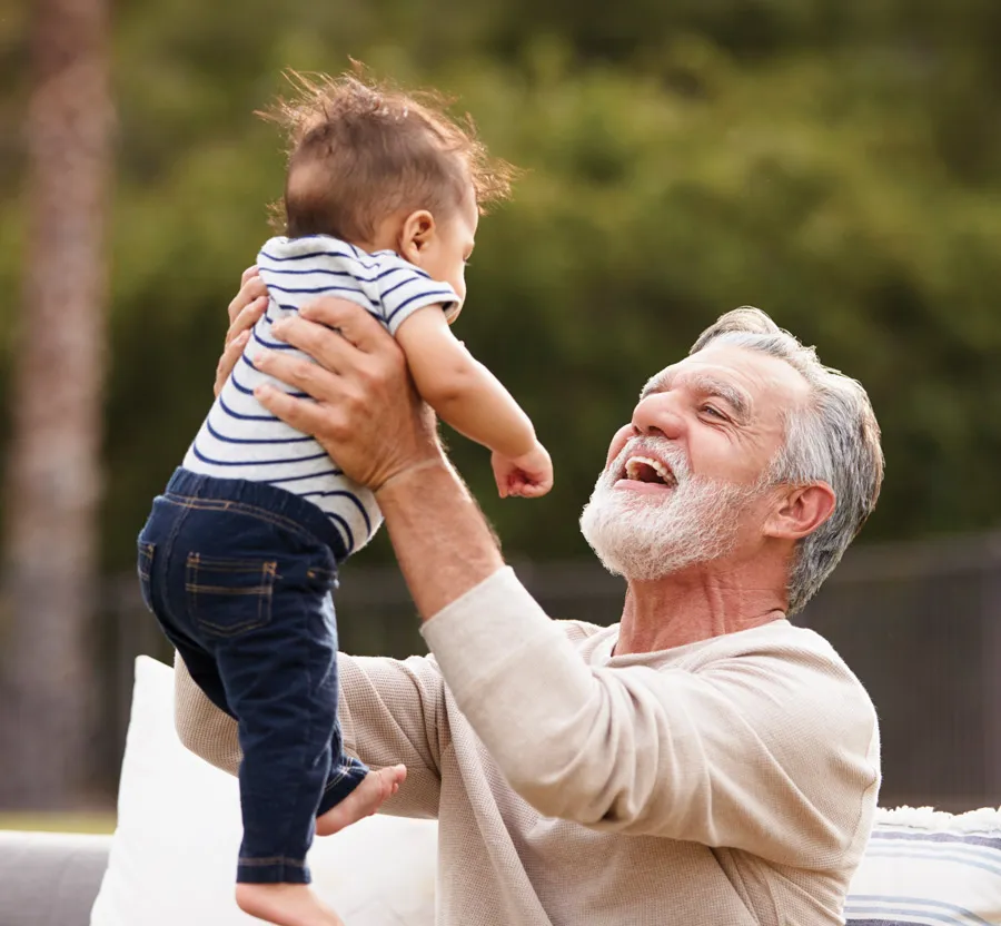 Joyful grandfather lifting his baby grandson outdoors, both smiling with a blurred green background.