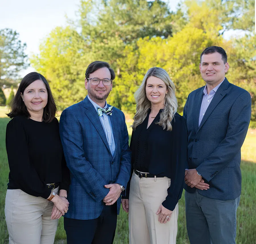 Four professionally dressed adults standing outside with trees and grass in the background smiling at the camera