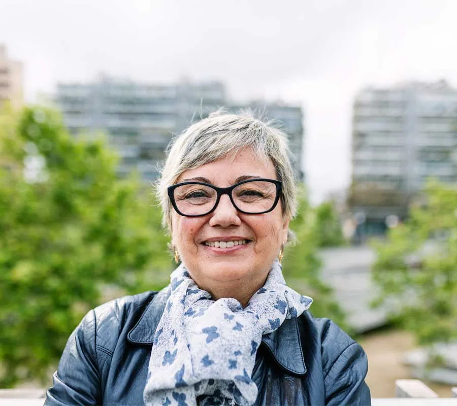 Smiling older woman with glasses wearing a patterned scarf and black leather jacket outdoors in urban park.