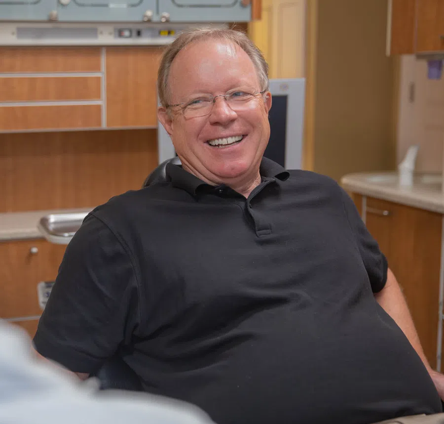 Smiling middle-aged man wearing glasses and a black polo shirt seated in a modern kitchen or office space.