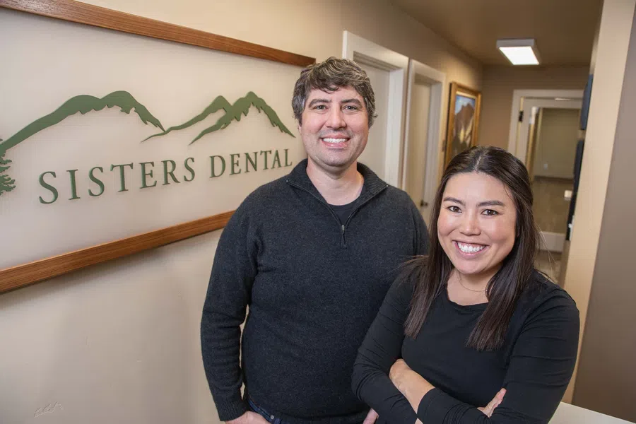 Two dental professionals smiling inside Sisters Dental office with logo and mountain design on wall.