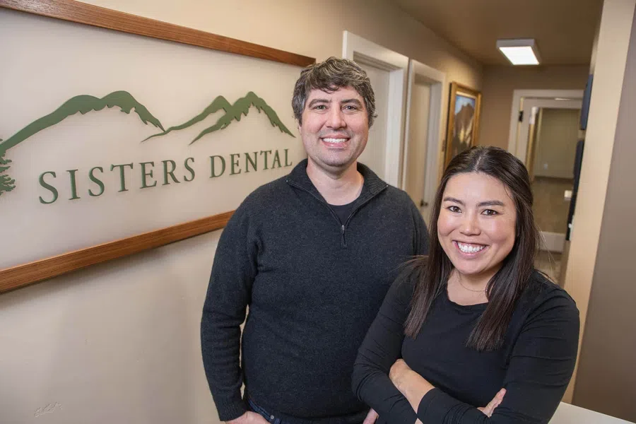 Two dental professionals smiling inside Sisters Dental office with logo and mountain design on wall.