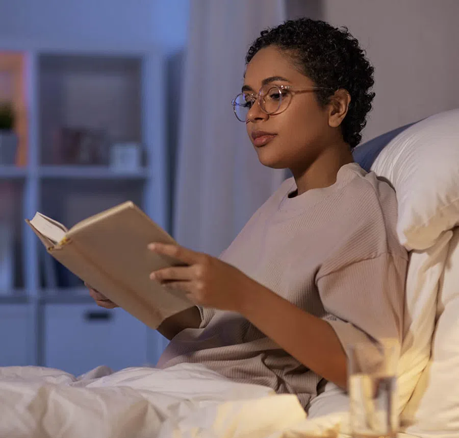 Young woman wearing glasses reading a book in bed at night with a glass of water nearby