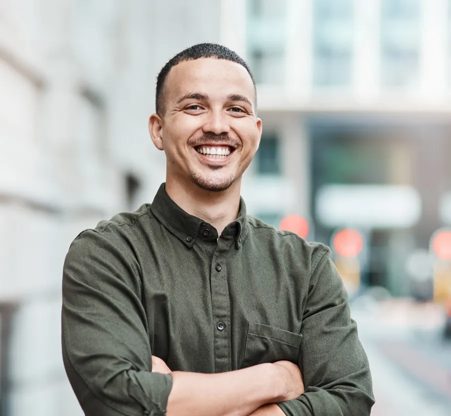 Smiling young man with crossed arms wearing olive green shirt standing outdoors in urban setting