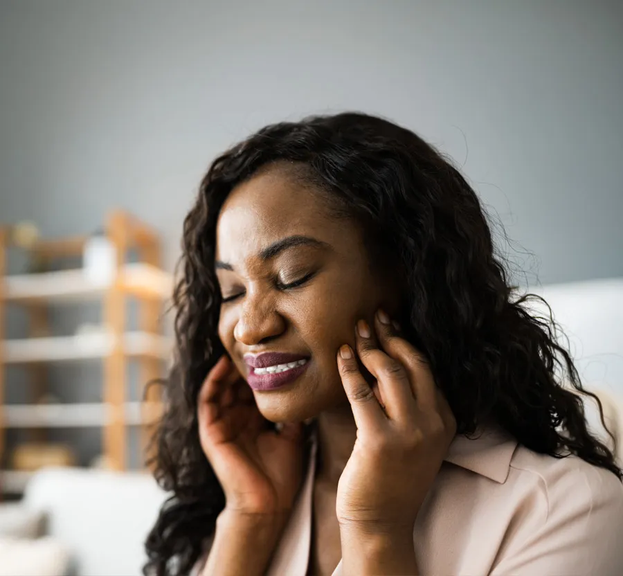 Woman with curly hair experiencing tooth pain and holding her jaw with eyes closed indoors.