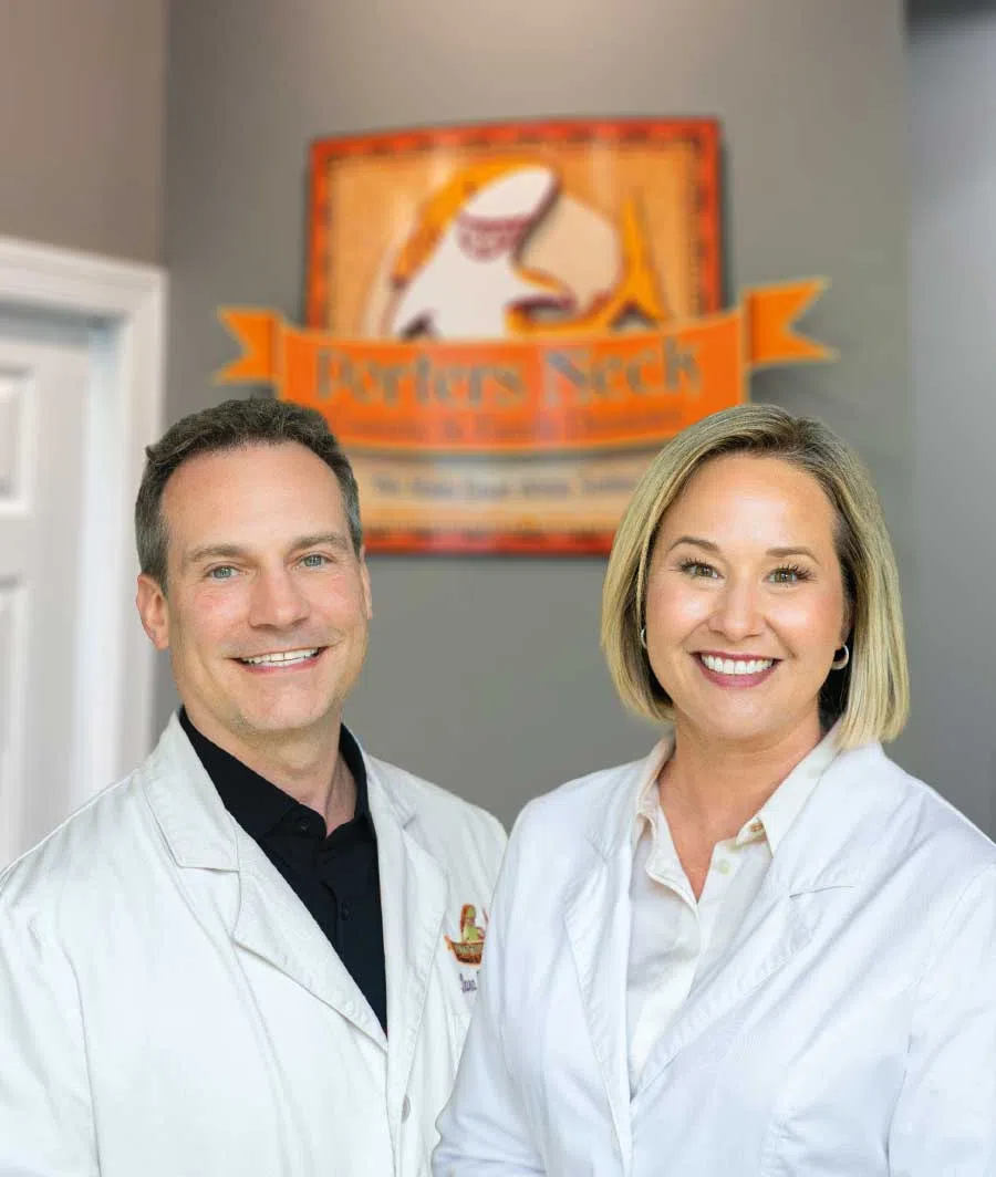 Smiling male and female medical professionals in white coats standing inside a clinic with an orange sign in the background.