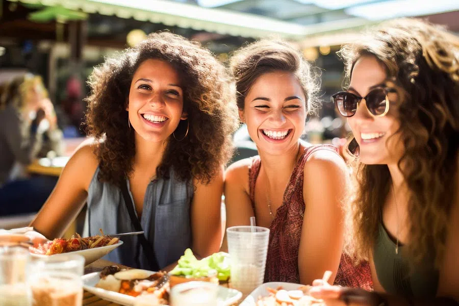 Three young women smiling and enjoying a meal together at an outdoor restaurant on a sunny day