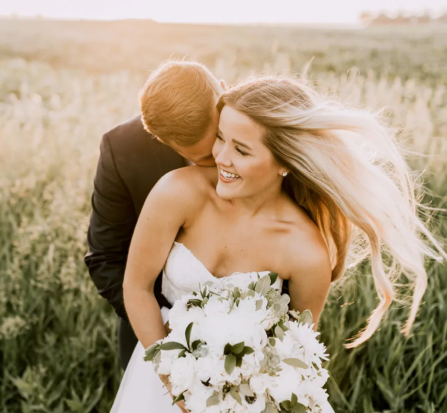 Joyful bride and groom embracing in a sunlit field with a white floral bouquet on their wedding day