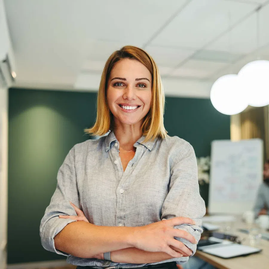 Smiling confident woman with crossed arms in modern office setting with blurred background and whiteboard.