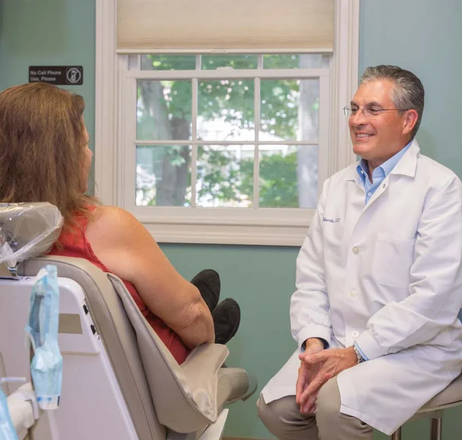 Smiling male doctor in white coat talking with female patient in medical office by window.