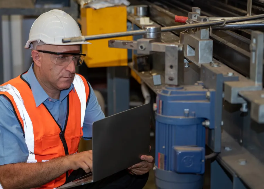 Factory worker in safety gear using laptop to monitor machinery operation in industrial setting