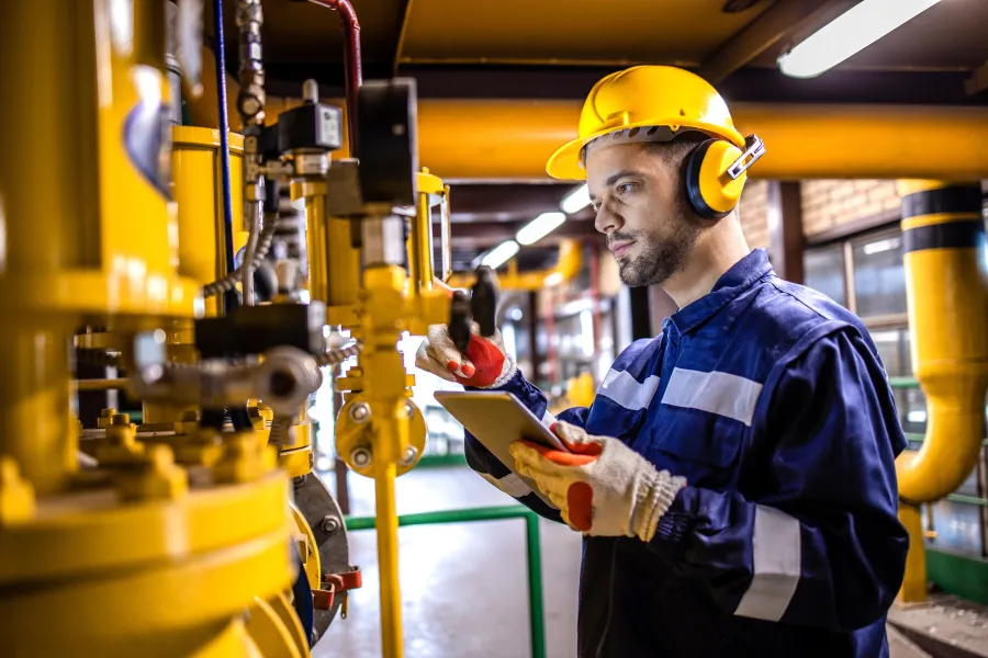 Industrial engineer in safety gear inspecting machinery and using tablet in a factory environment with yellow pipes.