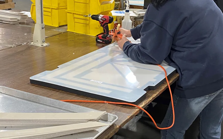 Worker using an air tool on a large white door panel in a workshop with yellow bins and tools.