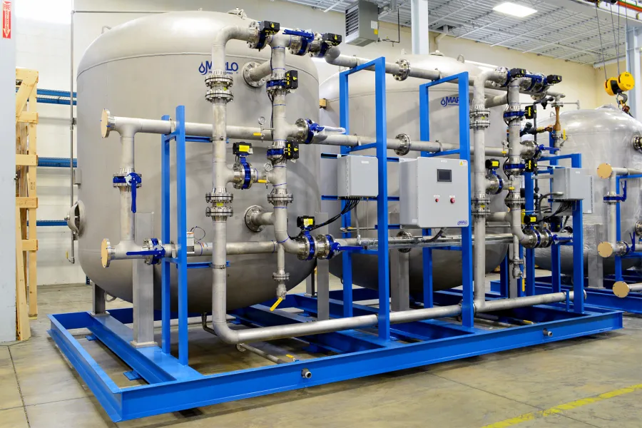 Industrial water treatment system with large metal tanks, pipes, valves, and control panels on a blue frame