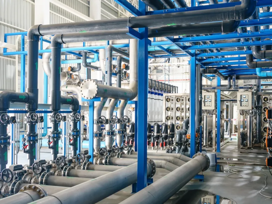 Industrial water treatment plant interior with pipes, valves, and filtration equipment under bright lighting.