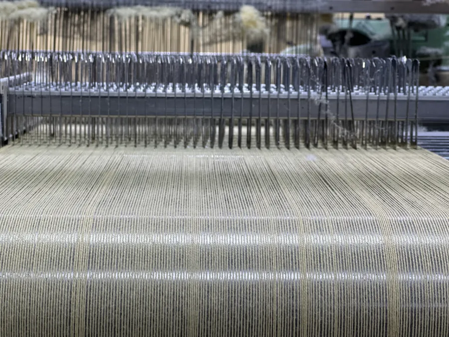 Close-up of industrial weaving machine producing beige fabric with parallel threads and metal needles.