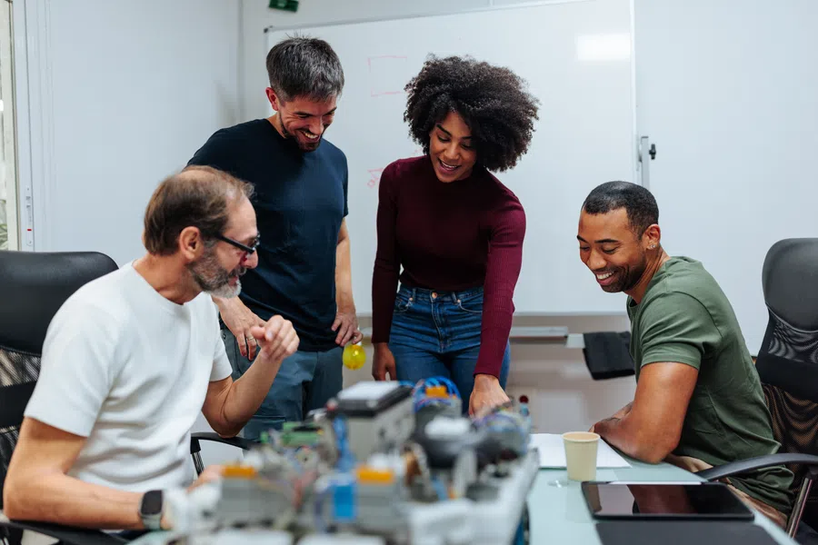 Four diverse coworkers collaborate over robotics project in modern office with whiteboard in background.