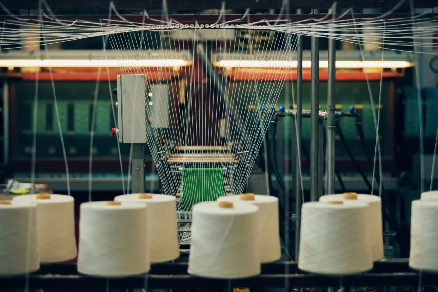 Spools of white thread feeding yarn into a weaving machine in a textile factory.