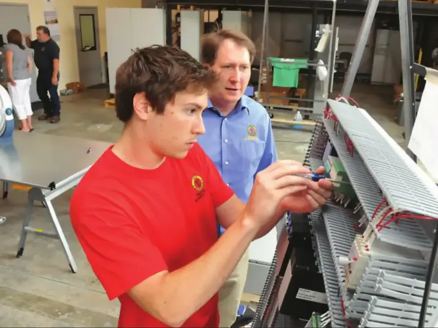 Young man in red shirt wiring an electrical panel while an instructor supervises in a workshop.