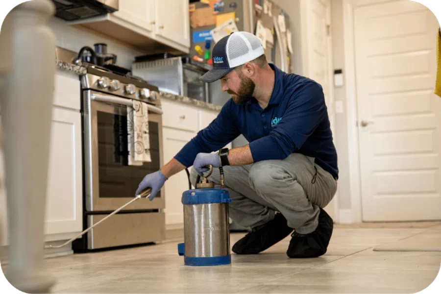 Pest control technician treating kitchen floor near stove with spray equipment wearing gloves and uniform