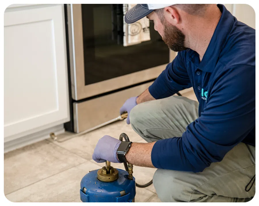 Technician using blue pest control equipment to treat area under kitchen oven wearing gloves and cap