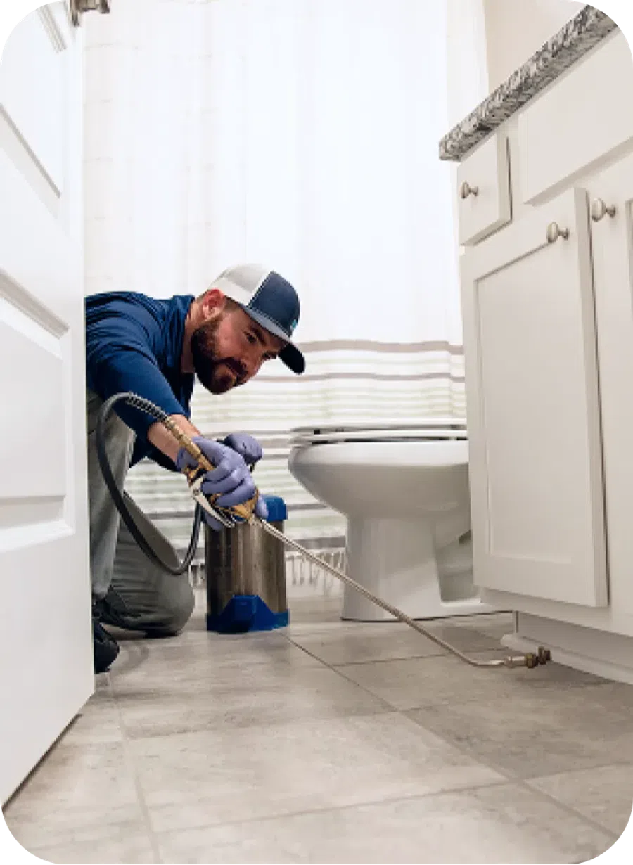 Pest control technician spraying insecticide around bathroom floor and cabinets with protective gloves.