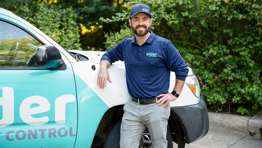Smiling pest control technician in company uniform leaning on branded service vehicle outdoors
