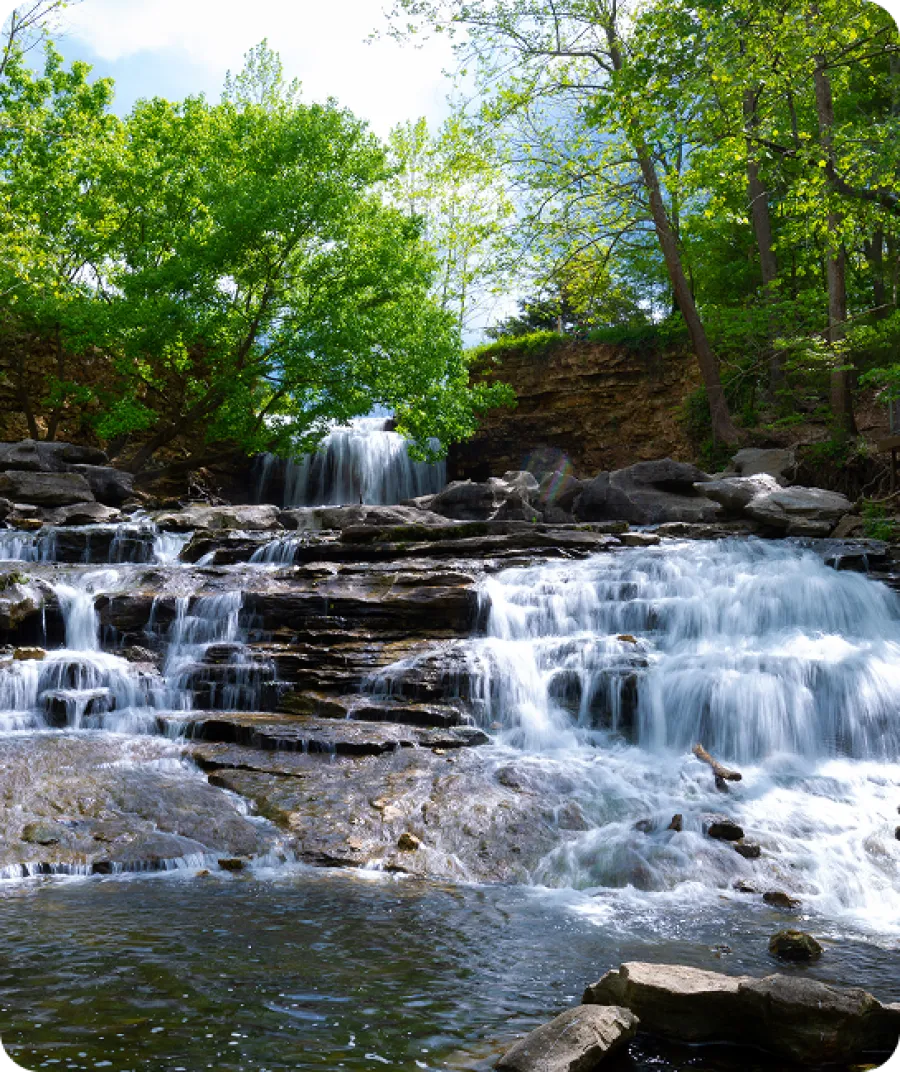 Serene waterfall cascading over rocks, surrounded by lush green trees and a clear blue sky.